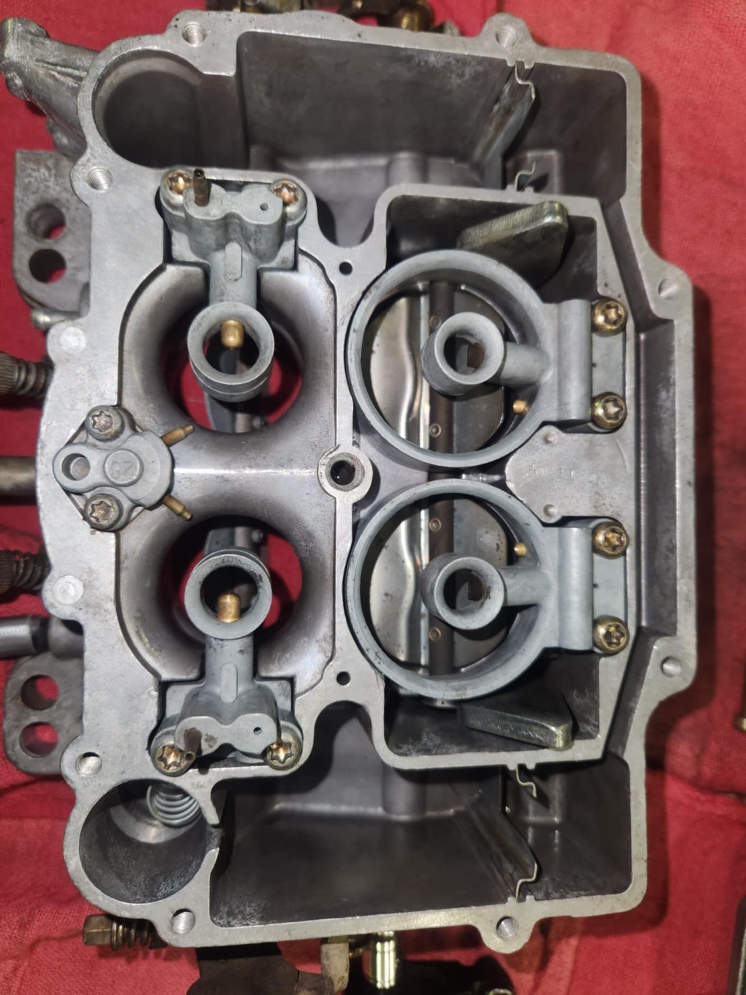 Top-down view of a four-barrel carburetor, showing the fuel bowls and venturi with grey and silver metal components.