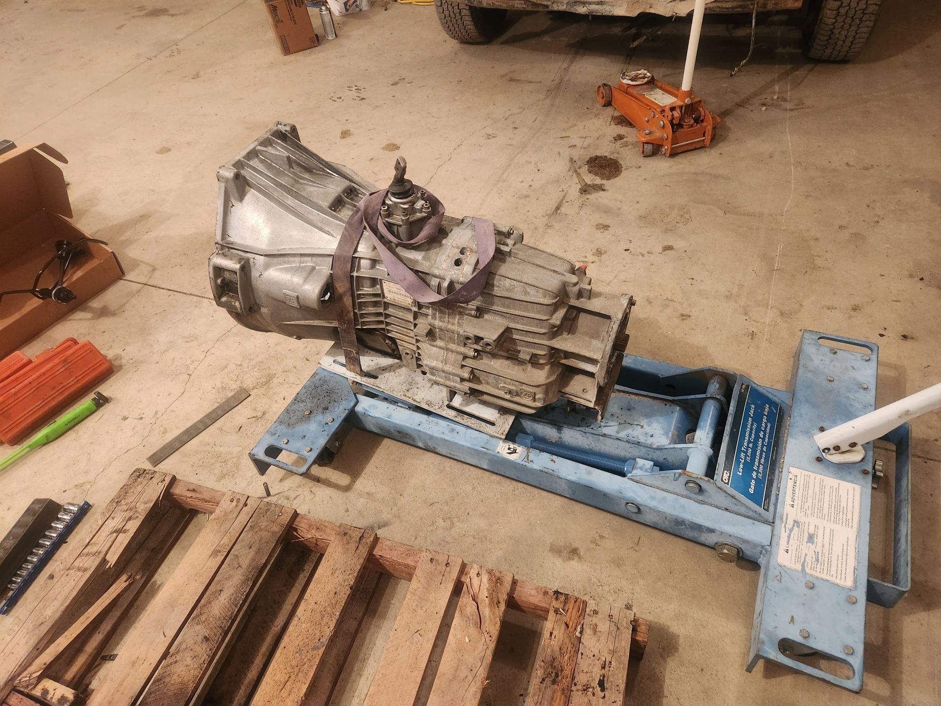 A large transmission on a blue transmission jack, sitting on a garage floor near a wooden pallet.