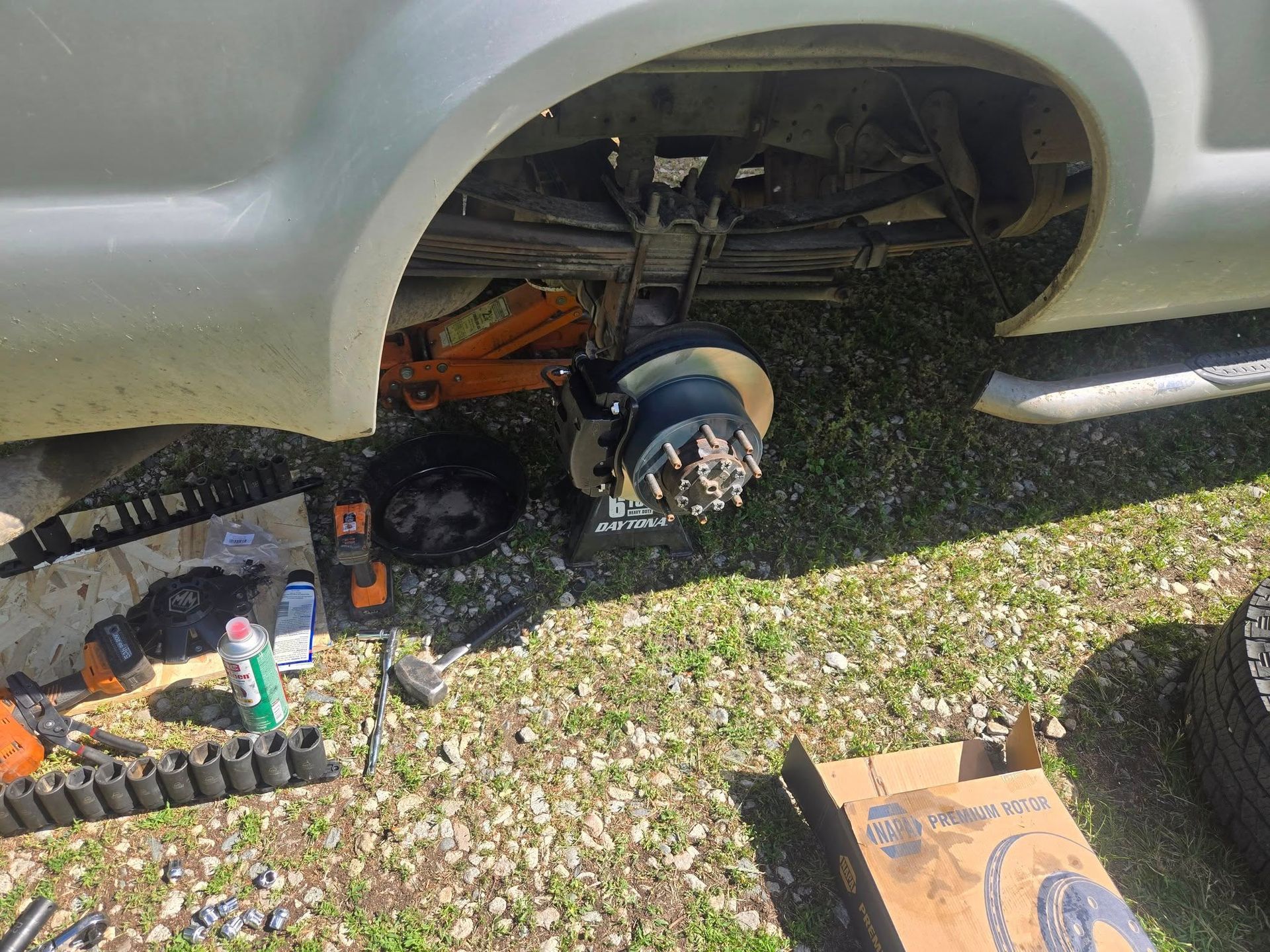 A truck's rear wheel is removed, revealing brake parts and suspension while it's lifted on a jack, tools scattered on the grass.