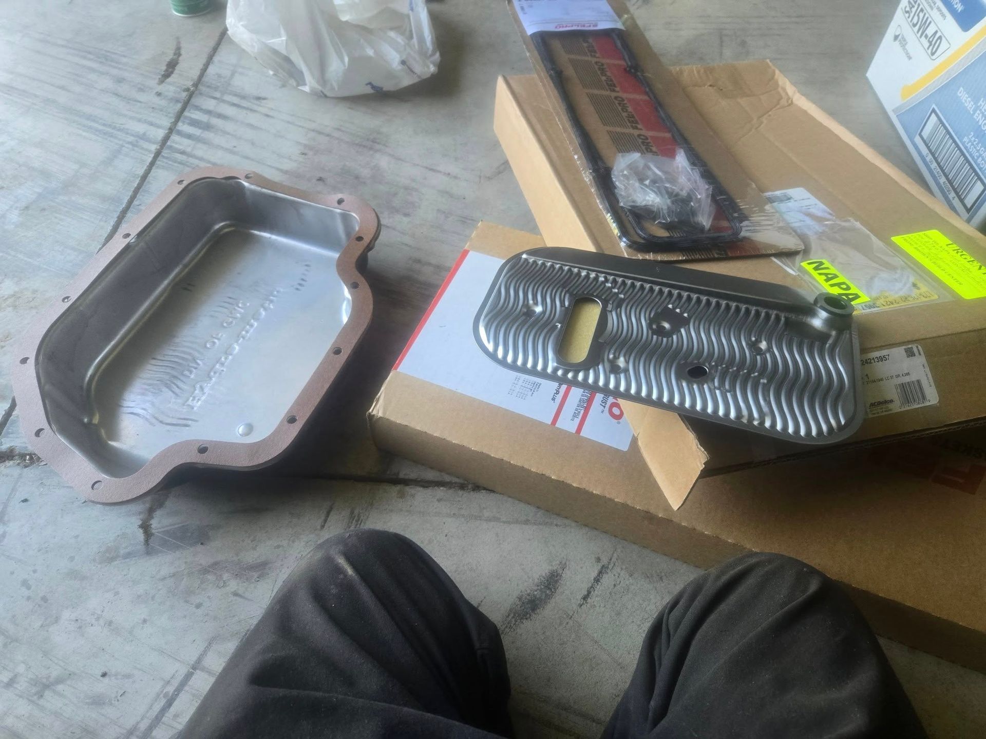 A car transmission pan, filter, and gasket on a garage floor, ready for installation.
