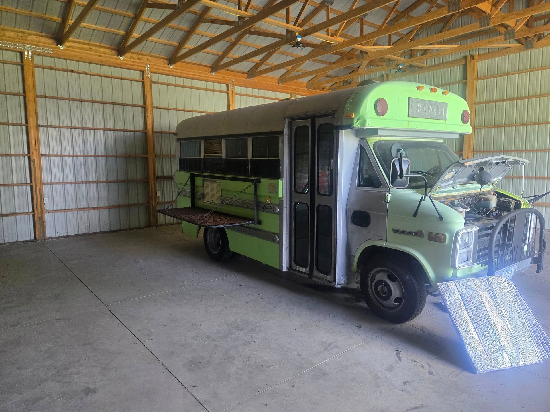 A repurposed school bus in a light green and gray color scheme, with its hood open, parked inside a large shed.