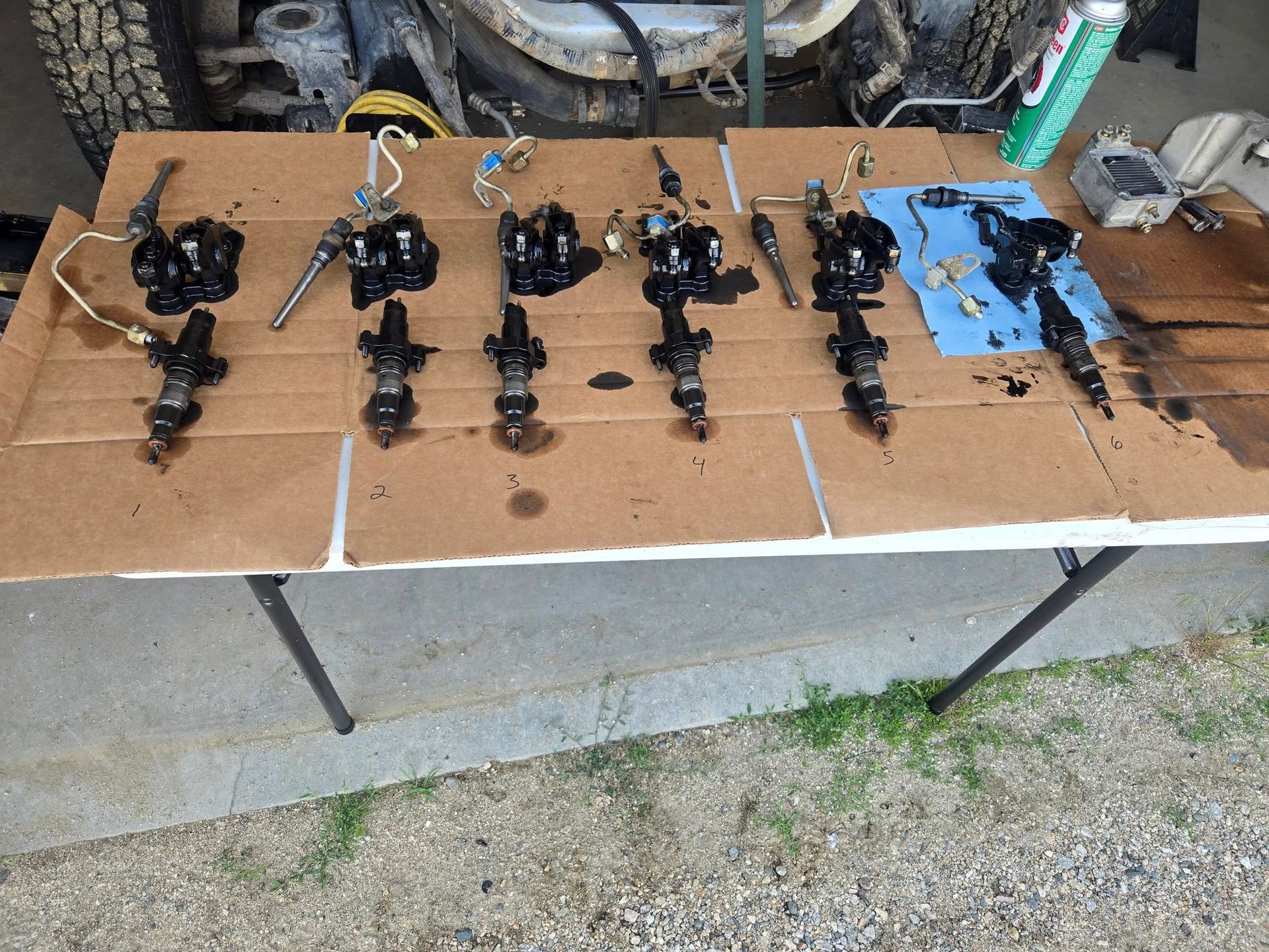 Diesel fuel injectors laid out on a cardboard-covered table, likely being serviced. Outdoor setting, with a vehicle visible in the background.