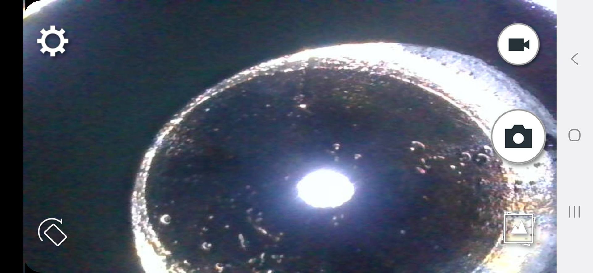 A close-up of a bright light source in the center of a dark, rounded shape, likely from a camera lens. Control icons are visible in the corners.