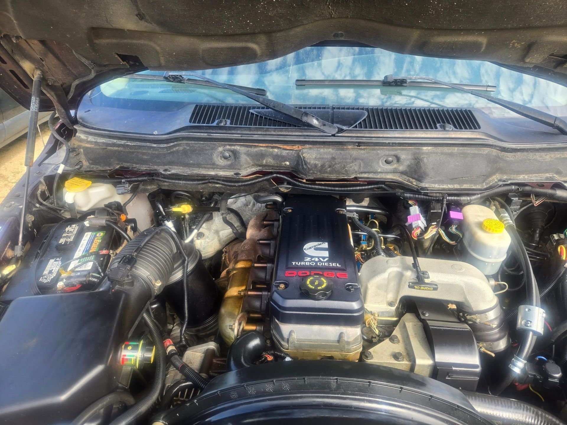 A black Dodge Ram engine bay with a Cummins diesel engine, showing various components, hoses, and wires. The hood is open and the windshield is visible.