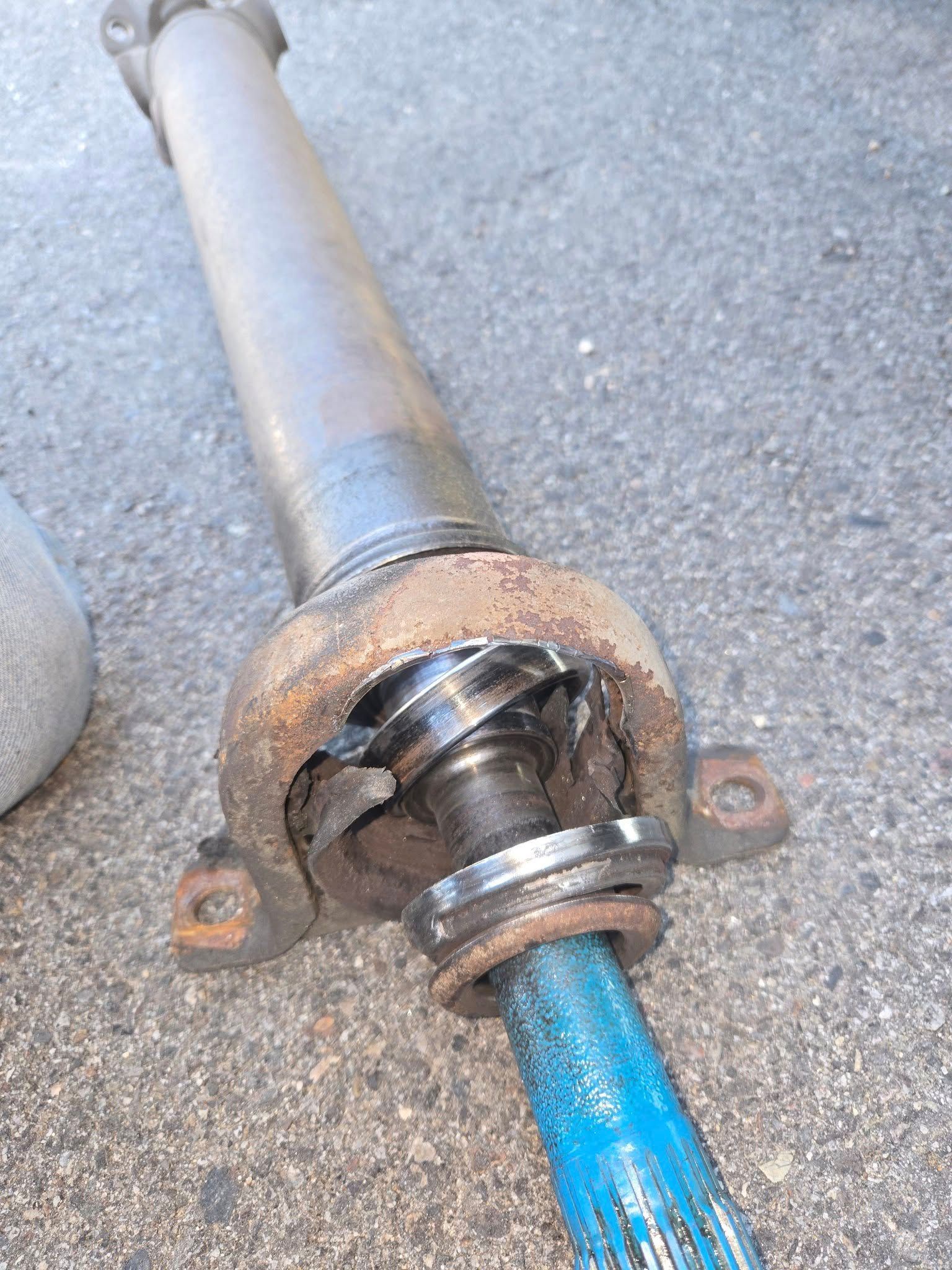 Damaged vehicle driveshaft on the ground. Metal components are rusty, with exposed blue shaft and broken bearing.