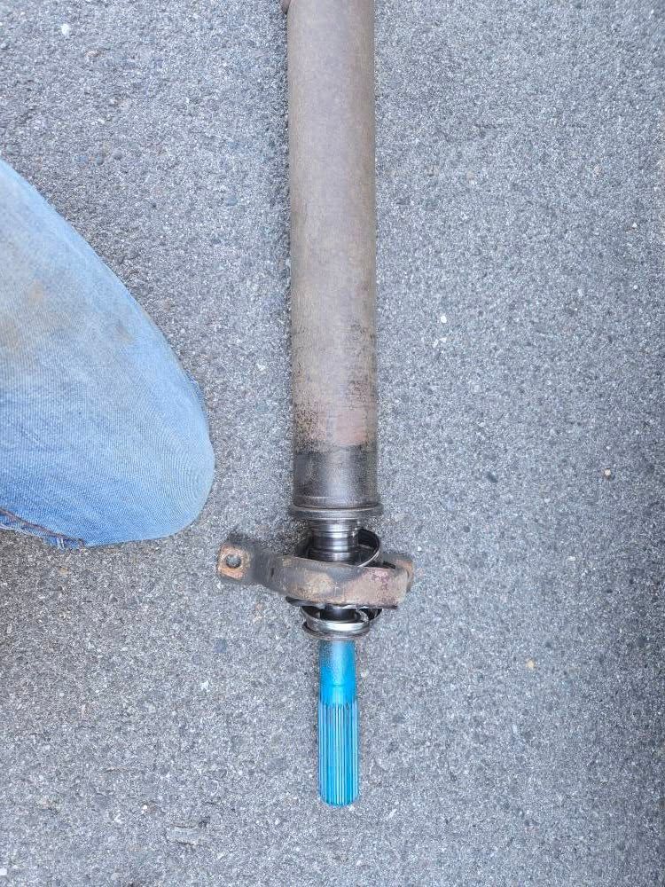 A rusty driveshaft, with a blue extension, lies on concrete. A foot in blue jeans is visible in the frame.