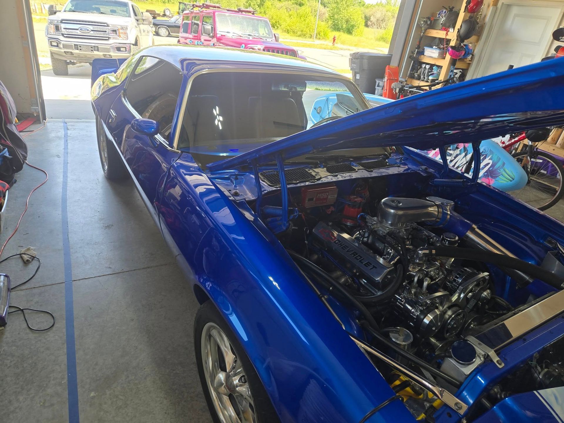 A shiny blue classic car with its hood open, parked in a garage with other vehicles visible outside.