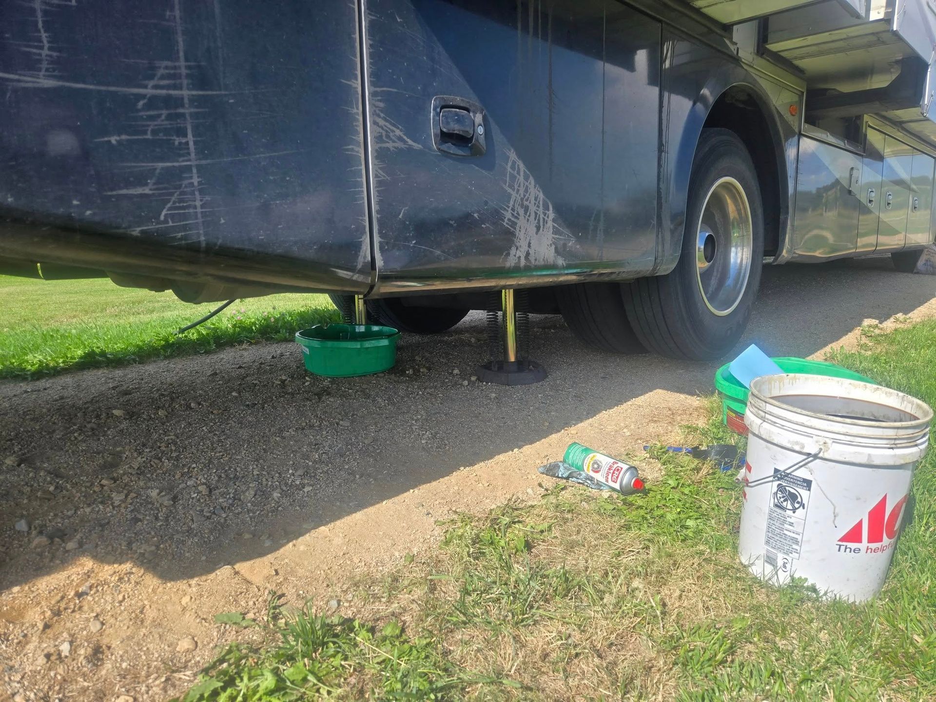 A dark RV is having an oil change. Oil drains into a green bowl on a gravel driveway, with buckets and supplies nearby.