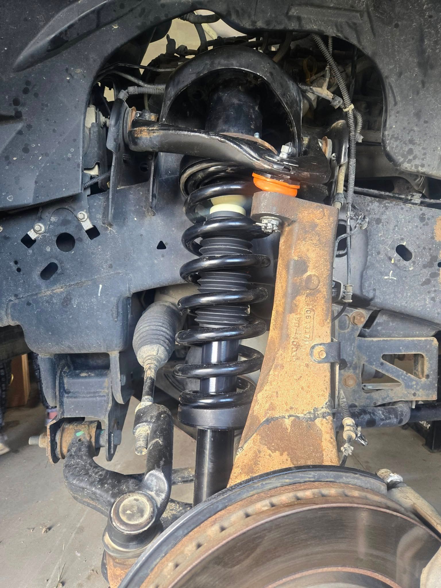 Car suspension system with black coilover shock absorber, orange accents, and rusty metal components.