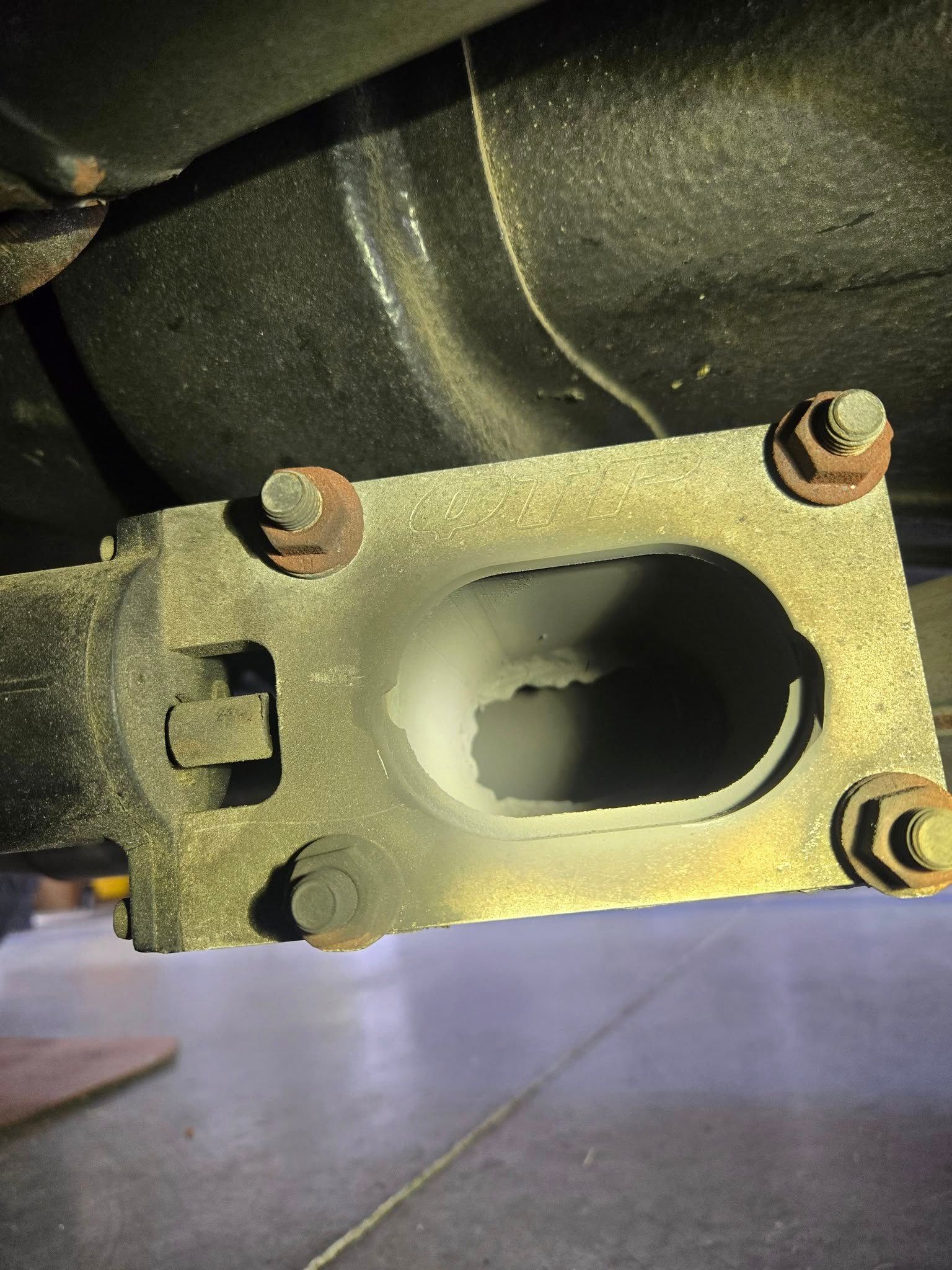 Underside view of a car's exhaust pipe flange, showing a rectangular opening and rusty bolts.