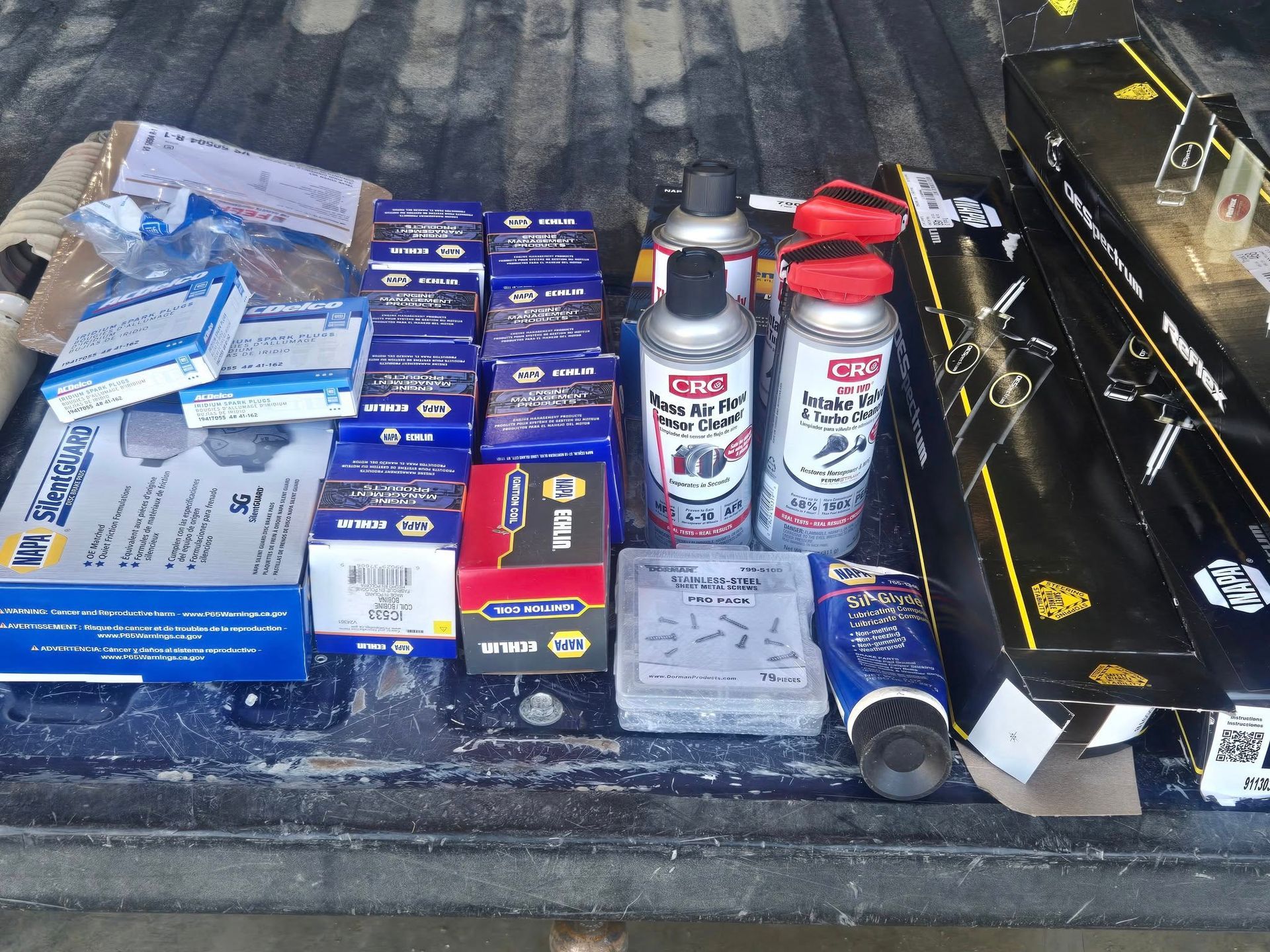 Various automotive parts and supplies are laid out in the bed of a truck, including boxes, cans, and tubes.