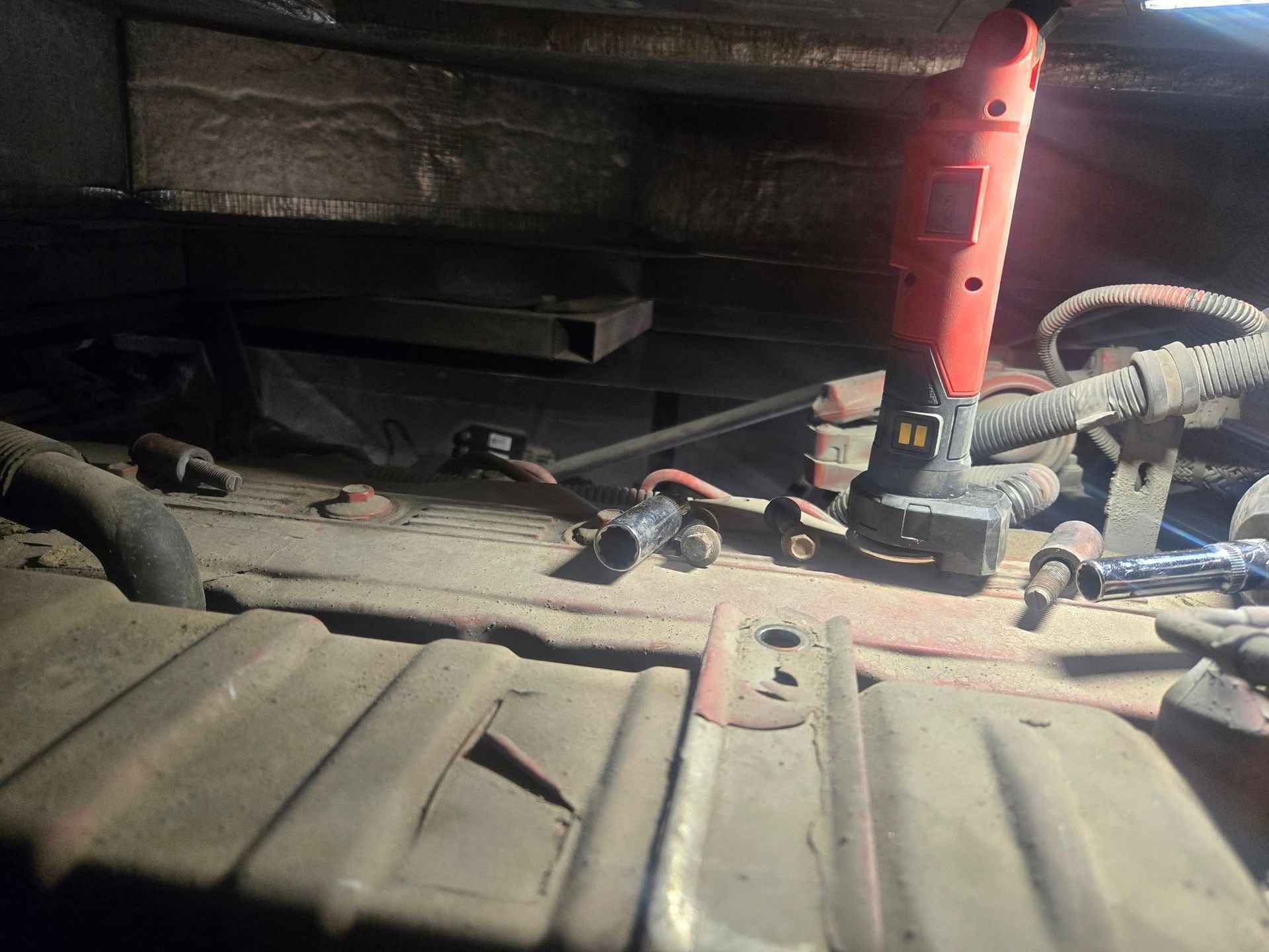 Inside a vehicle's engine bay, a red power tool illuminates a wiring harness and other components, viewed from a low angle.