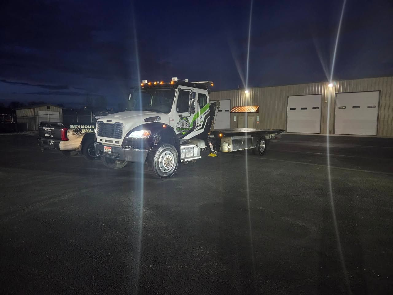 Tow truck parked on a wet lot at night beside an industrial building with garage doors.
