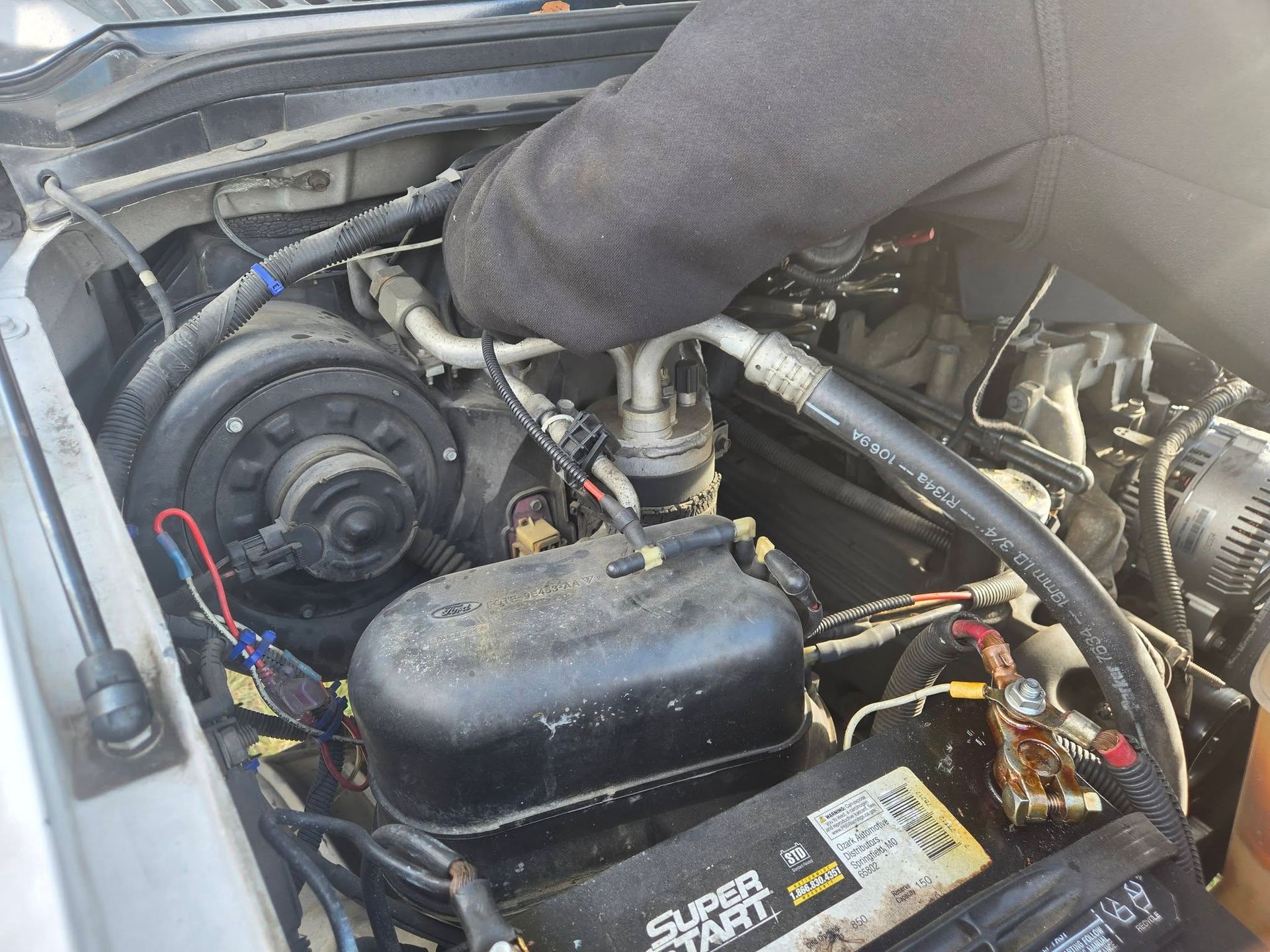 Car engine bay with a gloved hand working on hoses near the battery and belts