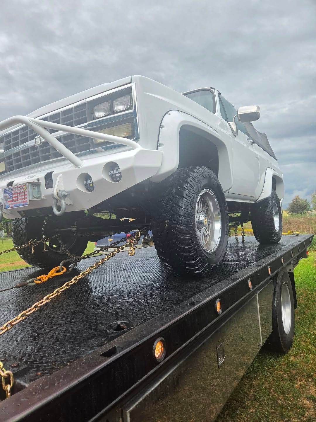 White lifted pickup truck on a black trailer in the rain, viewed from the rear side.
