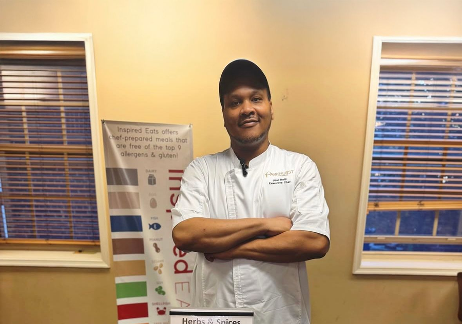 Chef at a food display. He stands with arms crossed, smiling. A banner and herbs are visible.
