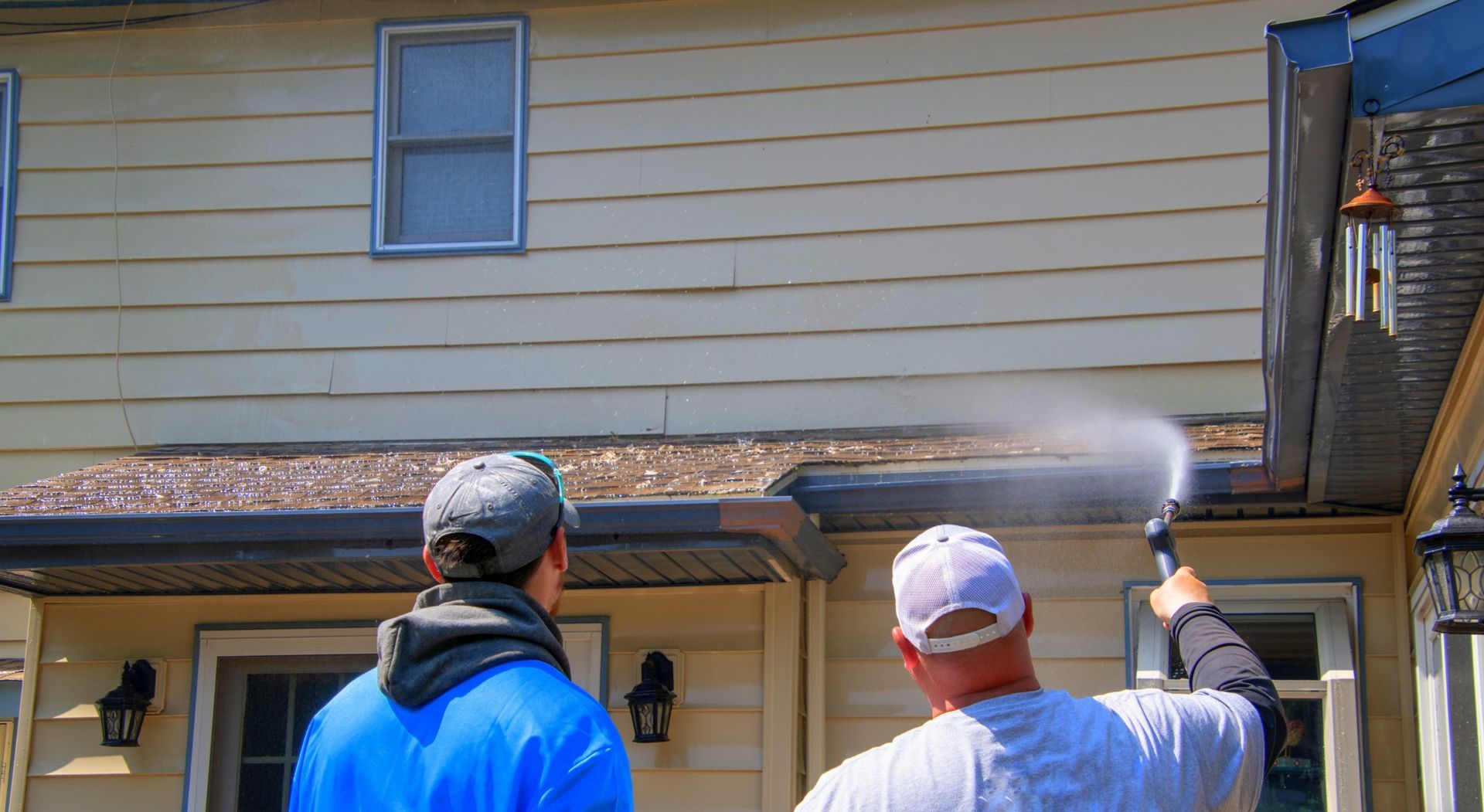 Two people pressure washing a house; one holds the sprayer, the other observes.