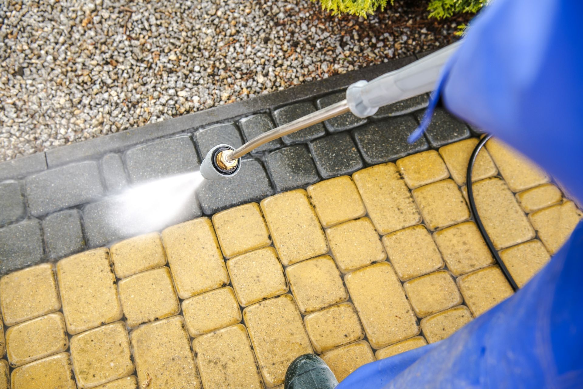 A pressure washer nozzle sprays water to clean a section of dark gray paving stones next to a yellow stone walkway.