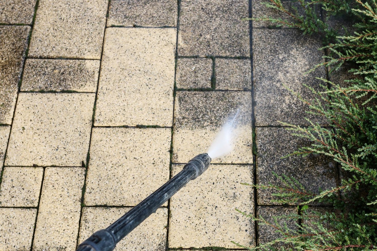 A person pressure washes a brick patio, cleaning the surface near a green bush.