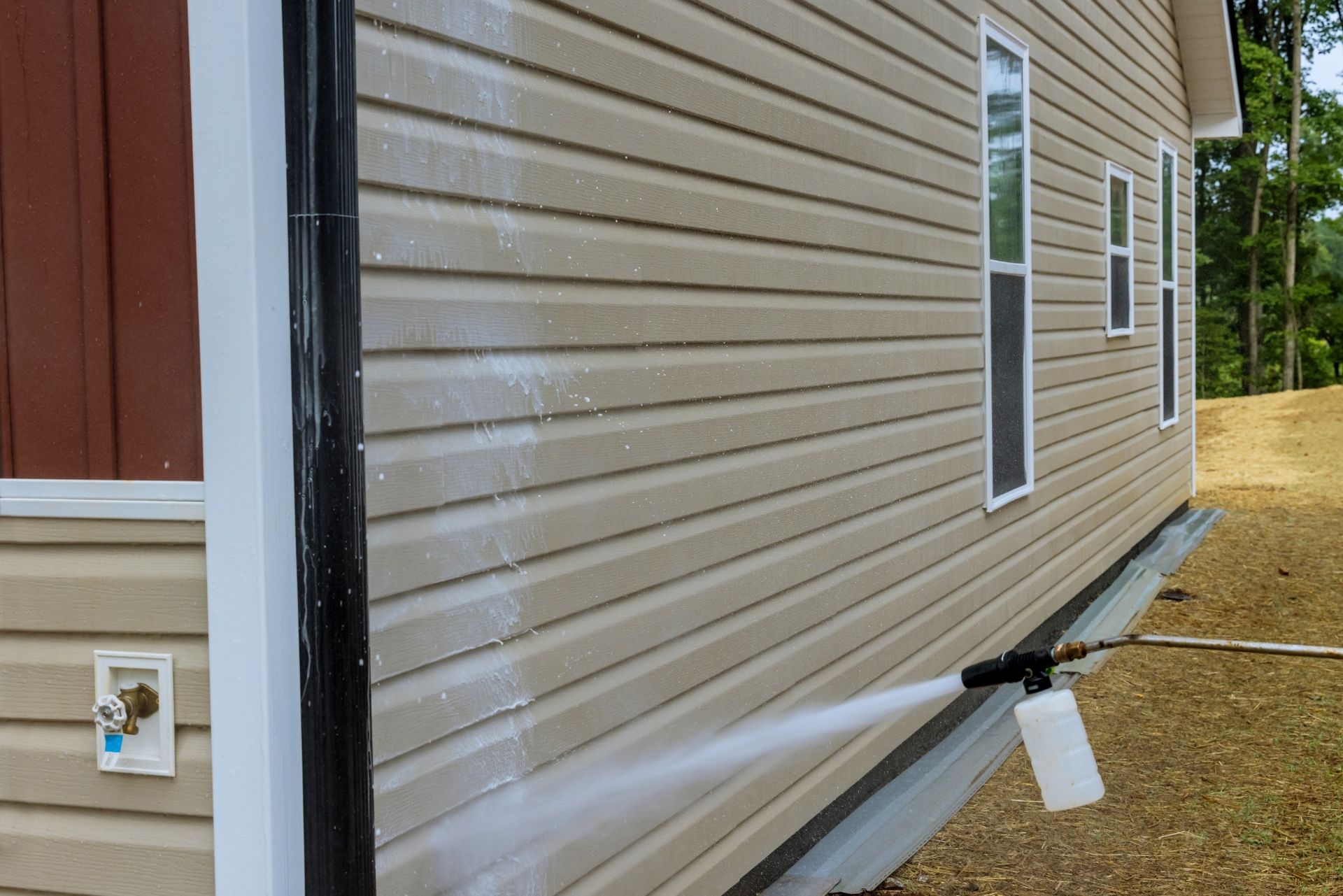 A person pressure washing the siding of a tan house. White foam coats the surface as water sprays.
