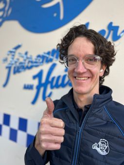 Een man met een bril en een zwart jasje zit lachend in de auto.