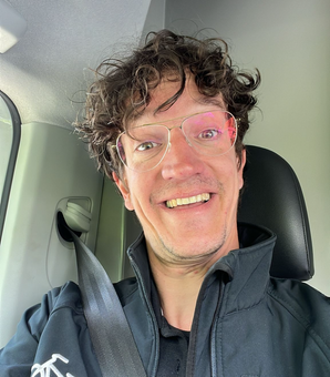 Een man met een bril en een zwart jasje zit lachend in de auto.