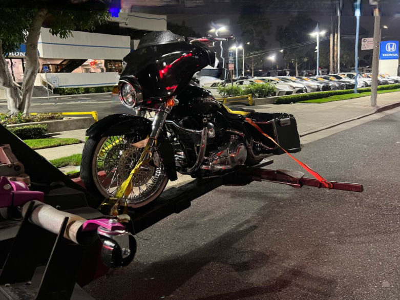 Black Harley-Davidson motorcycle secured on a tow truck at night. Bright lights, parked cars in background.