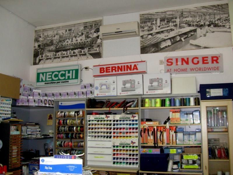 Interno di un laboratorio di cucito con marchi come Necchi, Bernina e Singer, oltre a fili, materiali e foto storiche della fabbrica.