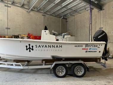 A white Savannah Expeditions boat on a trailer inside a large garage, featuring a black Mercury outboard motor.