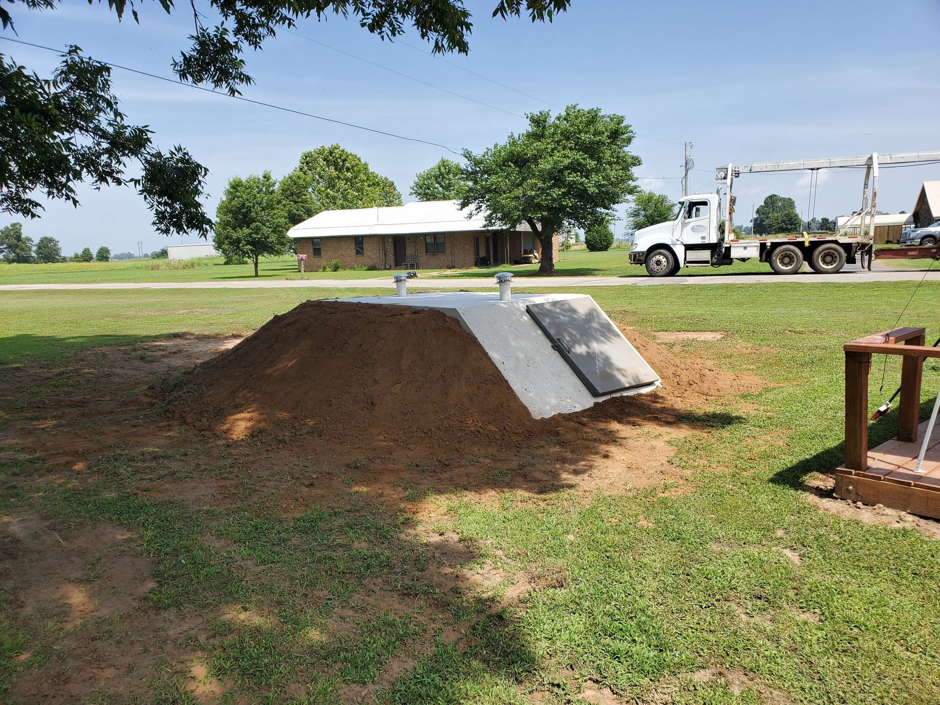 A concrete storm shelter partially buried in dirt, with a truck nearby and a house in the background.