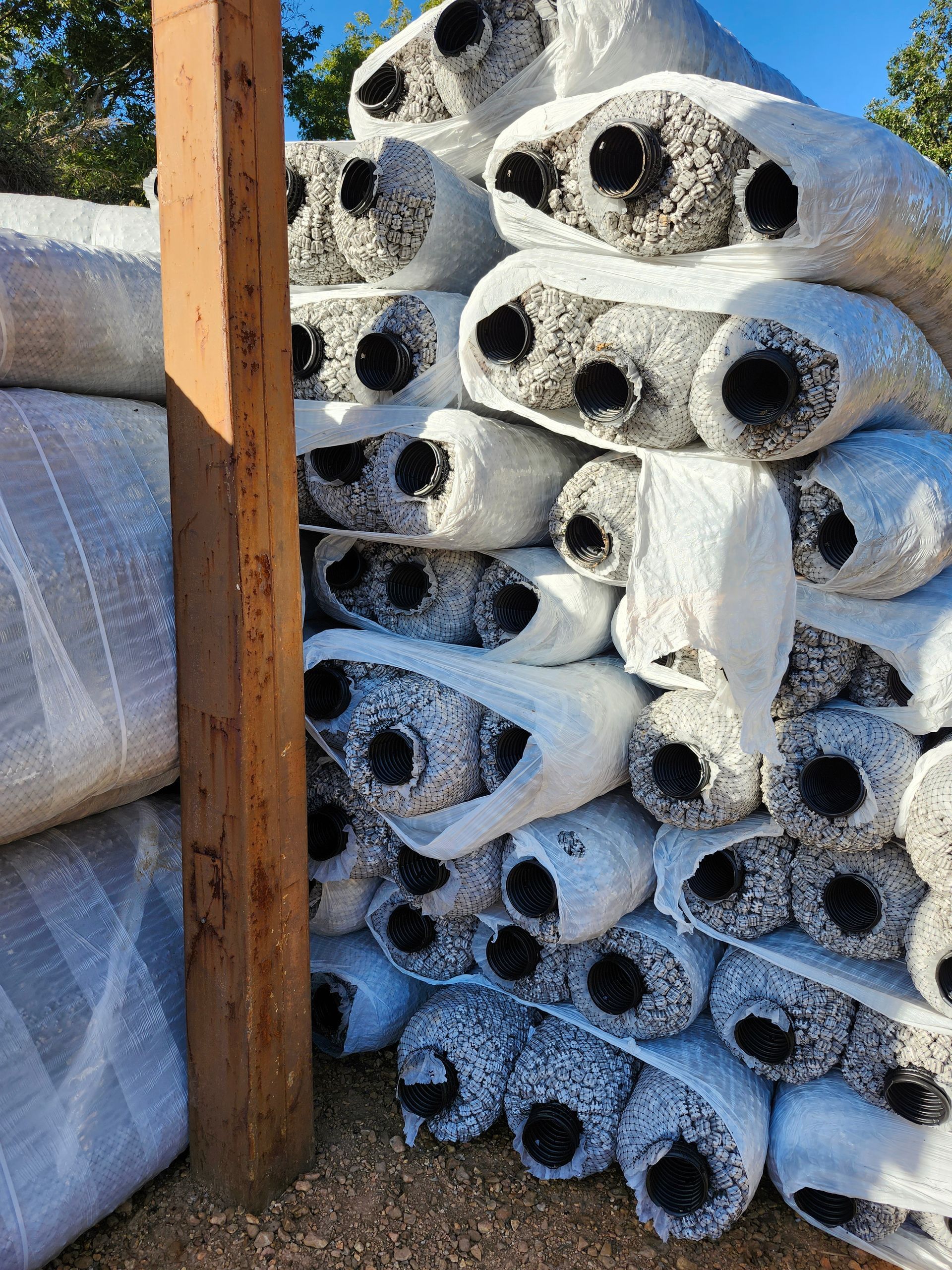 Stack of rolled, wrapped landscaping fabric. A wooden post is in the foreground.