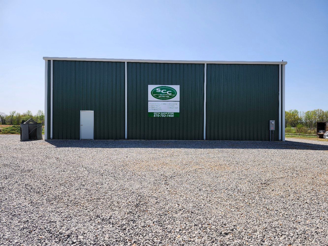 Green metal building with a sign on a gravel lot under a blue sky.