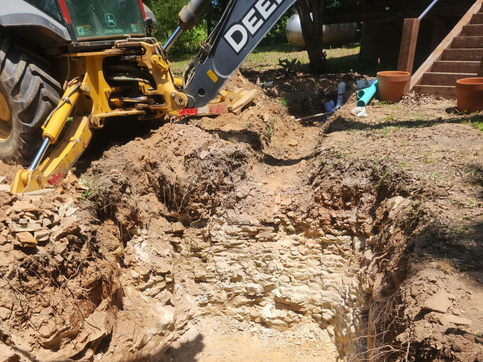Yellow backhoe digging in dirt near a wooden deck and potted plants.