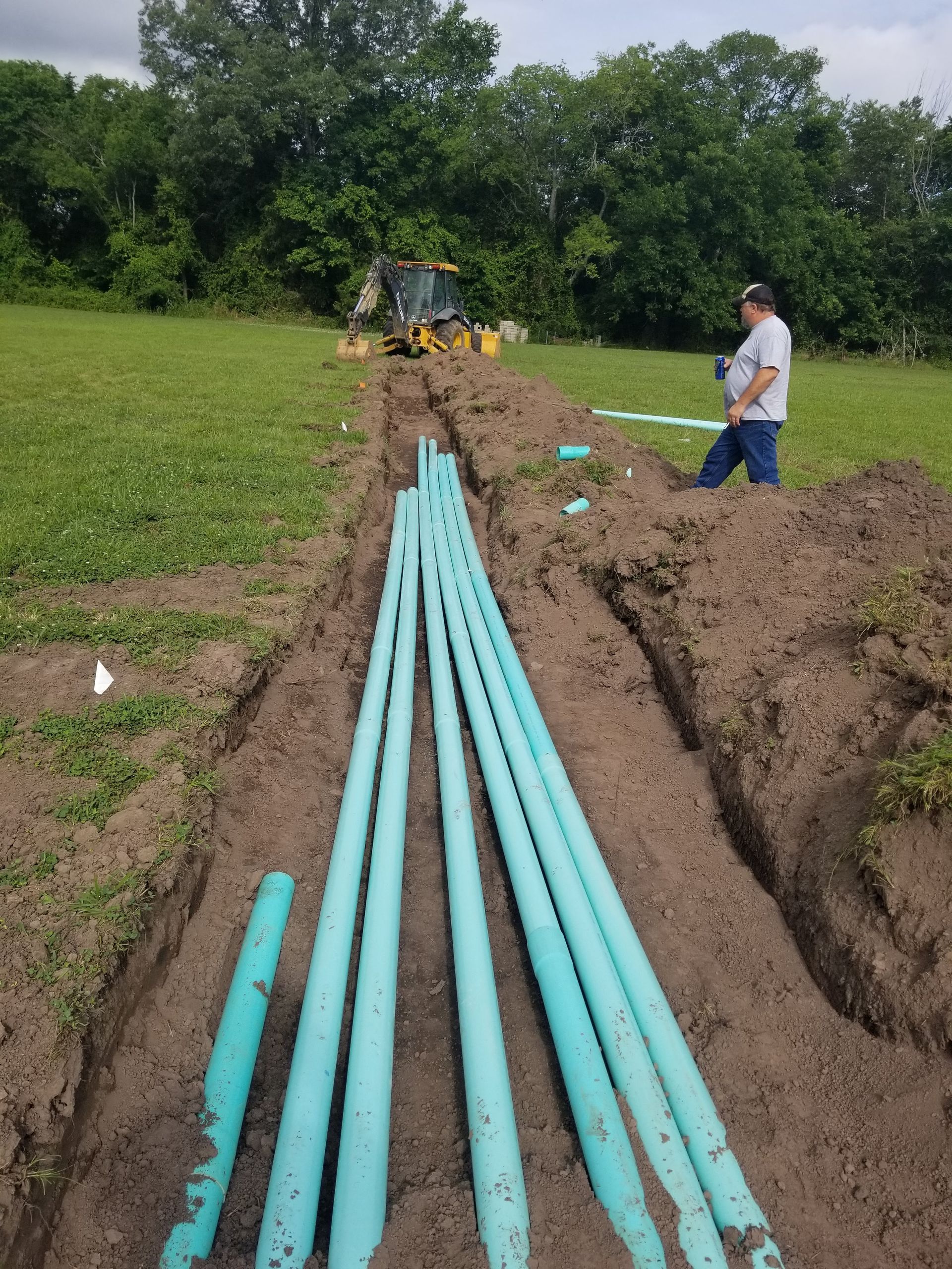 Trenches with turquoise pipes are being installed in a grassy field.