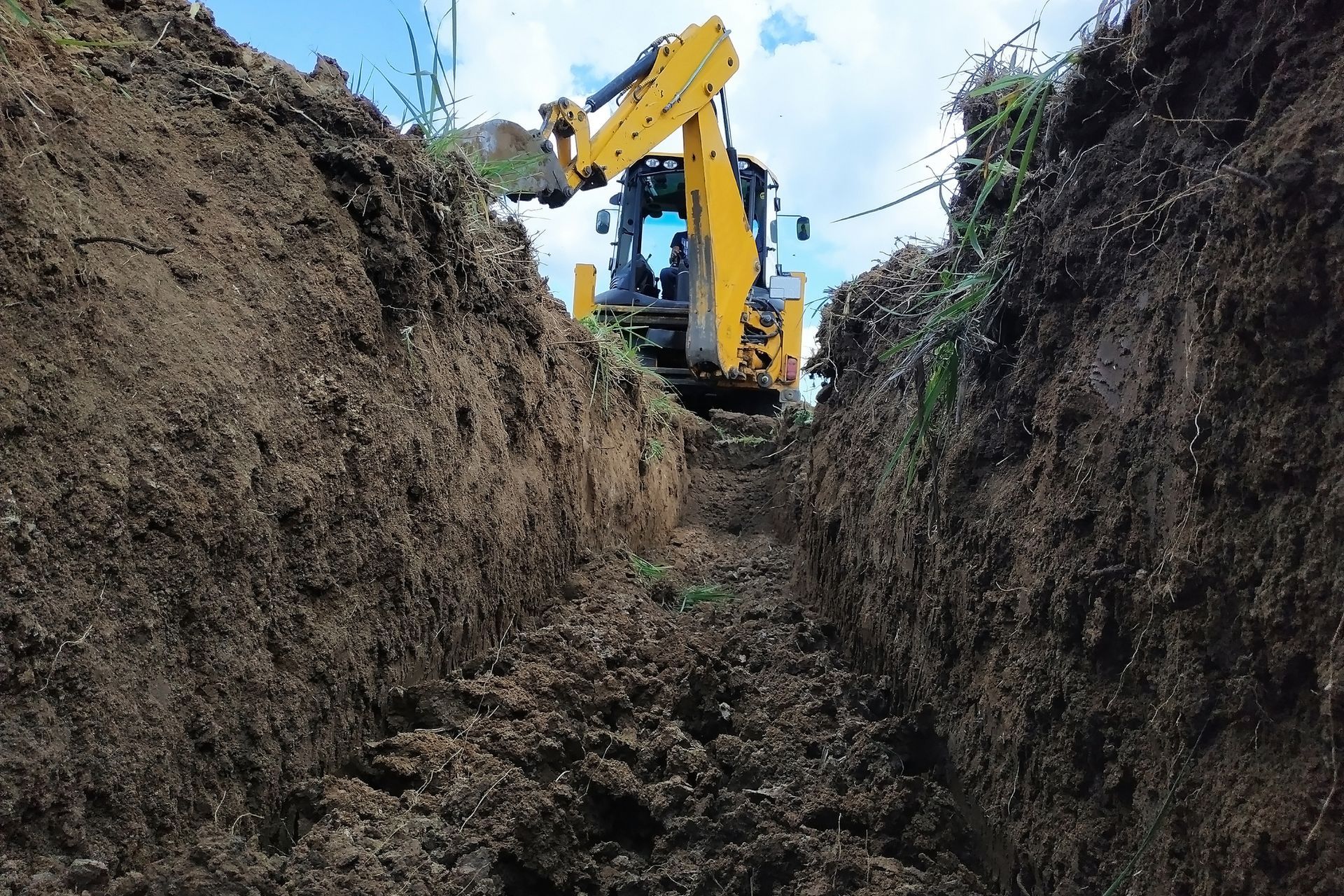 Yellow excavator digging a trench in the earth, against a cloudy sky.