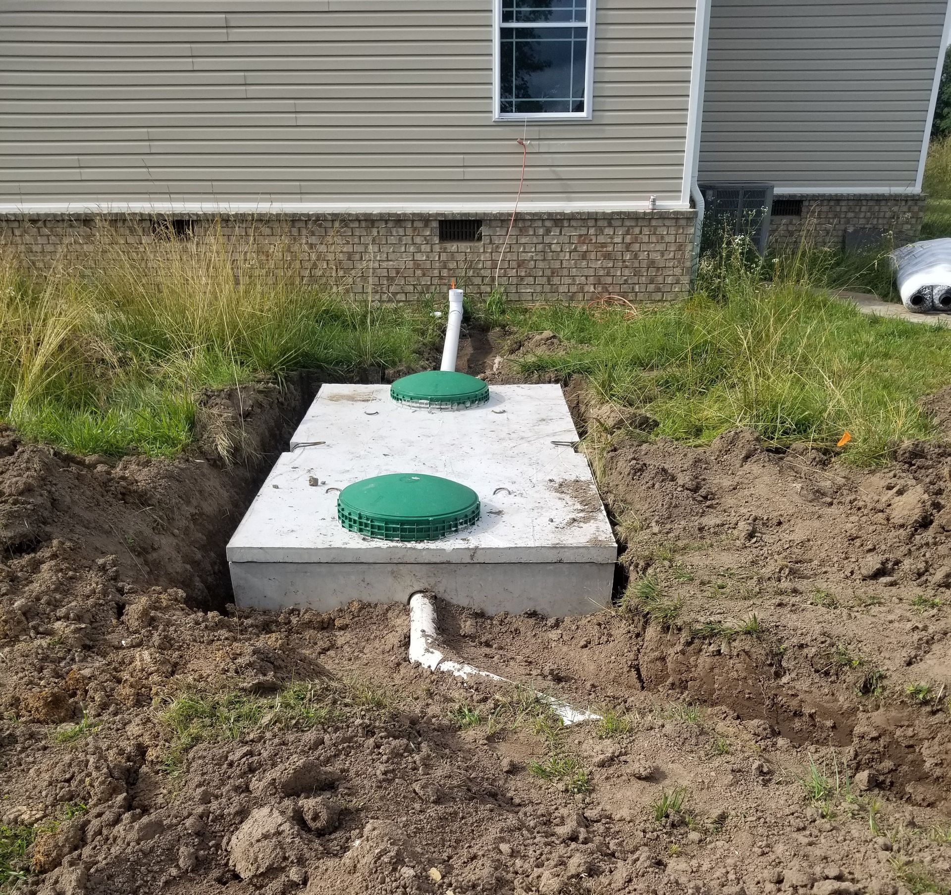 A rectangular concrete septic tank with two green circular lids, partially buried in the ground beside a residential home.