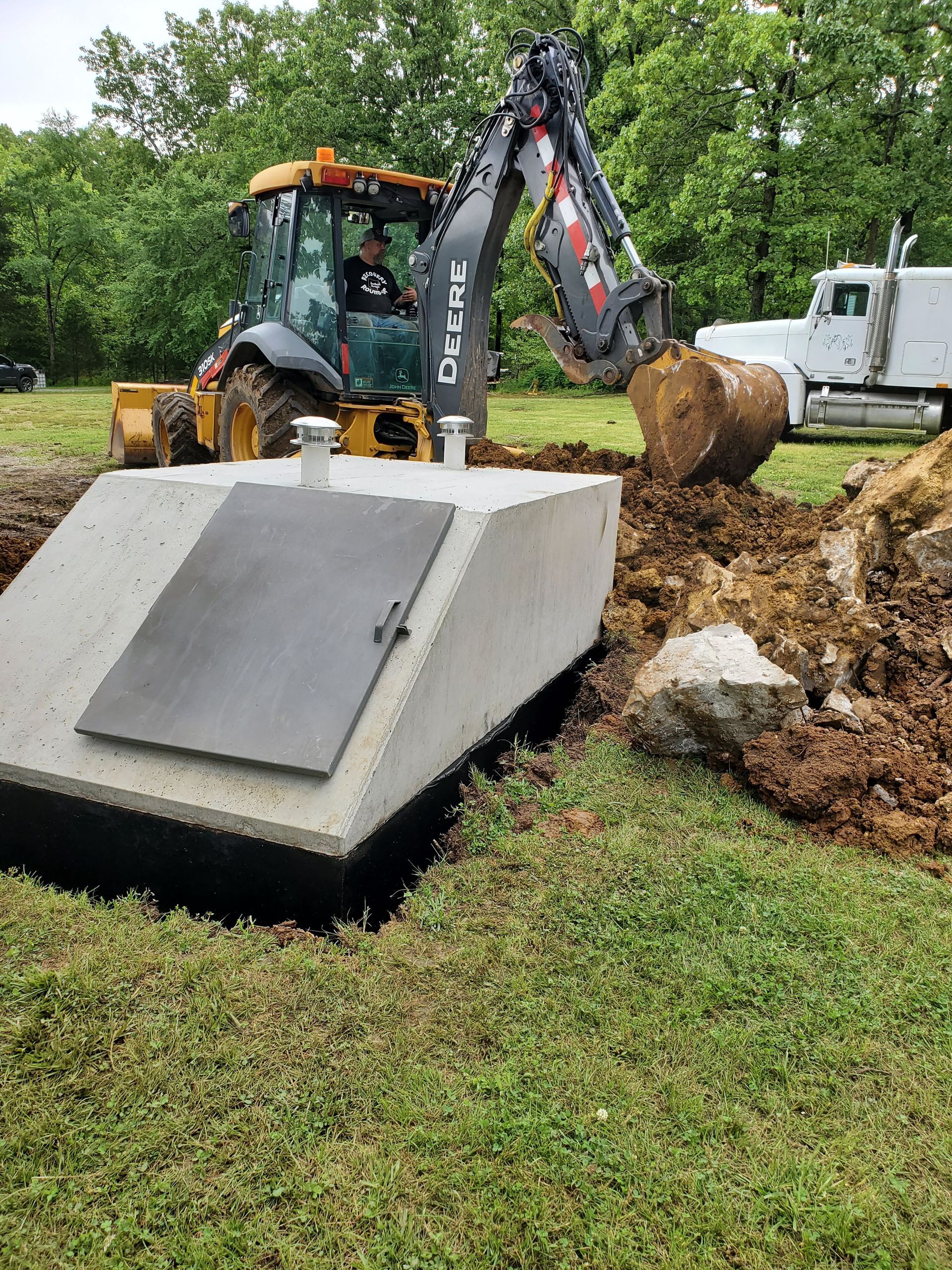 A yellow John Deere backhoe digs into the ground next to a concrete storm shelter on a grassy, wooded site.