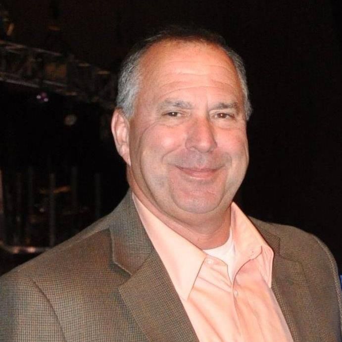 Man in a brown blazer and coral shirt smiles, posing against a dark background.