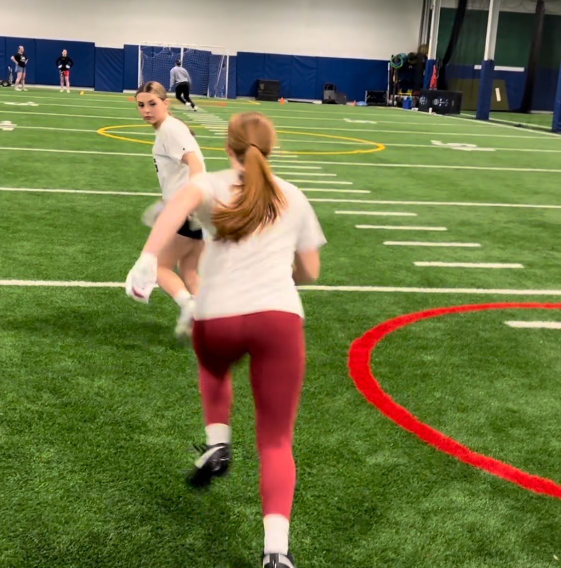 Two women running on a green indoor field, one in burgundy leggings, white shirt, and ponytail.