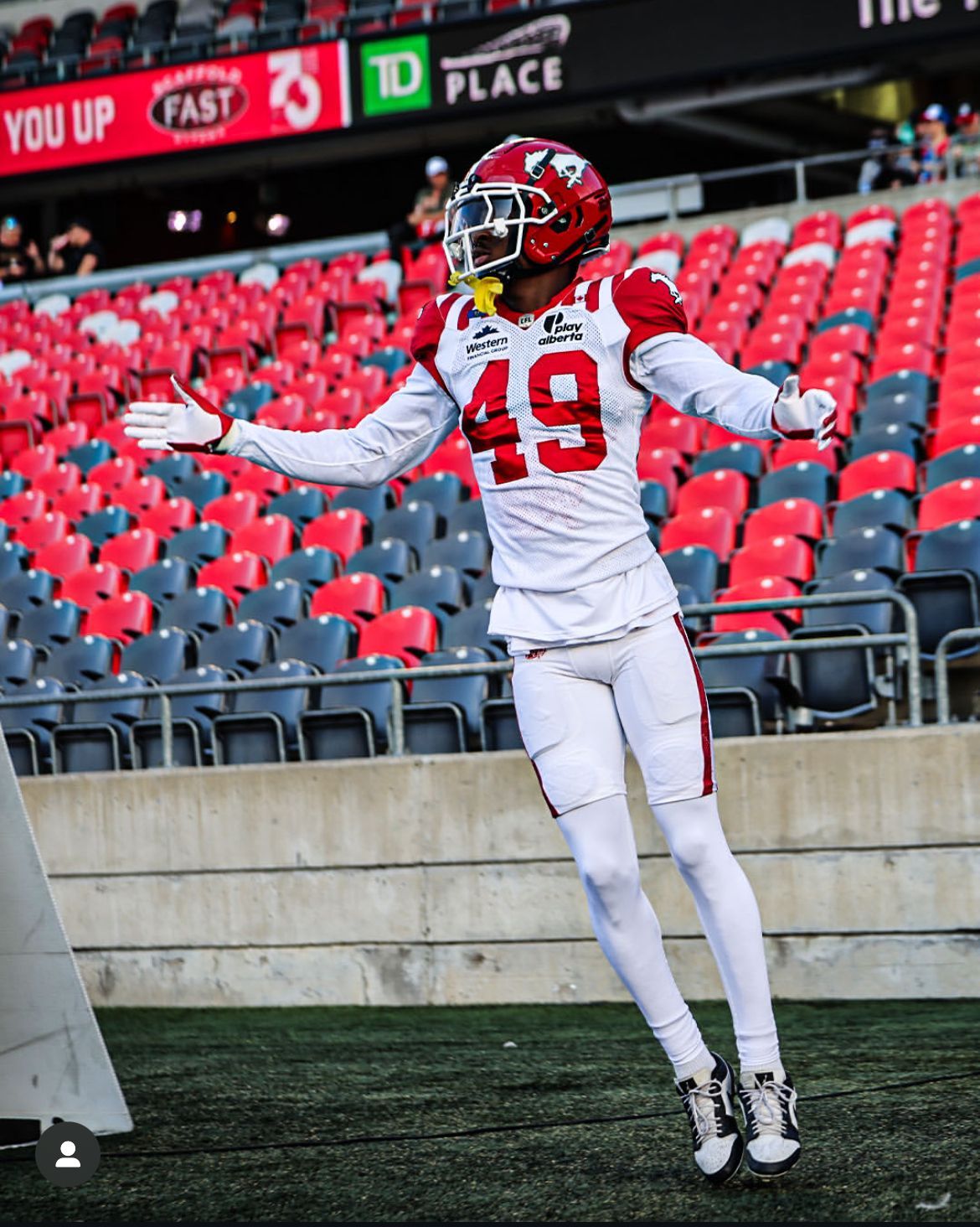 Football player in white uniform with red accents, number 49, leaps on a field in front of red stadium seats.