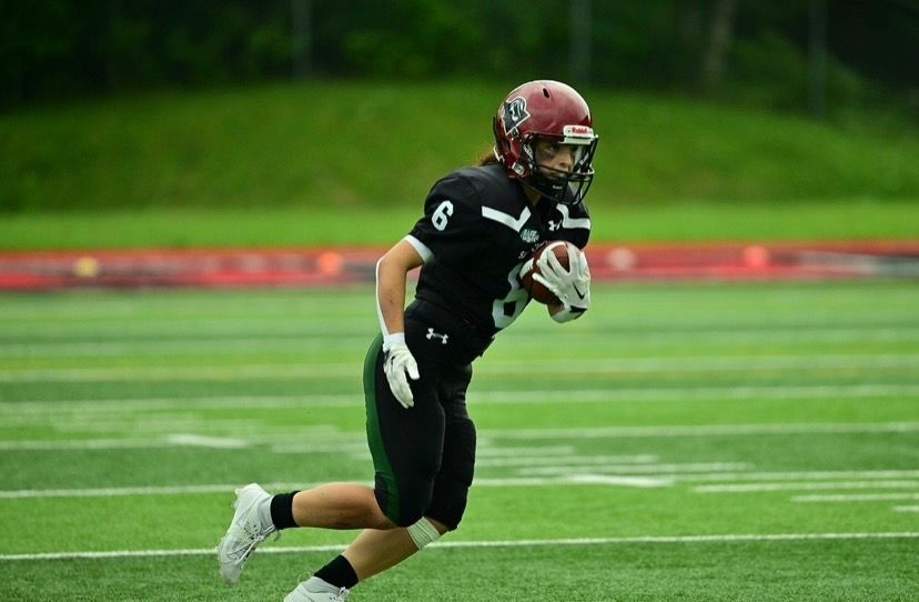 Football player in black uniform with number 6, running on a green field, holding a football.