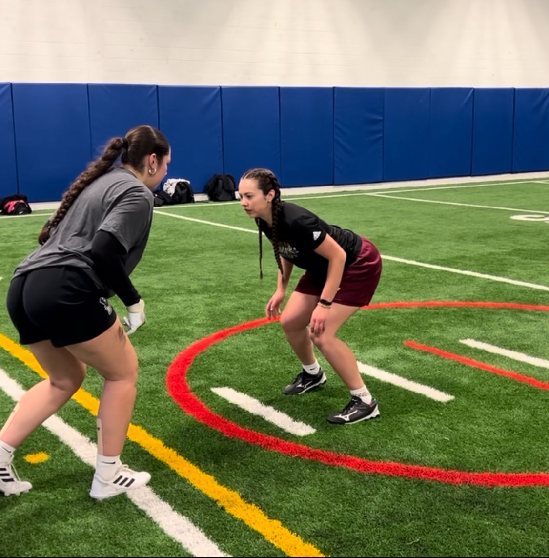 Two people on a turf field face each other in athletic stance, one with white gloves. Indoor setting.