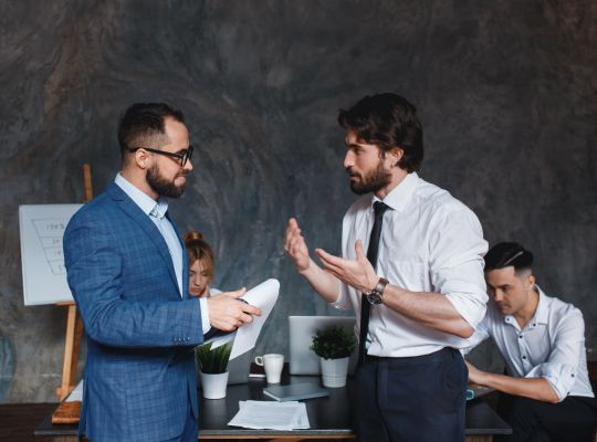 Two men are standing next to each other in an office talking to each other.