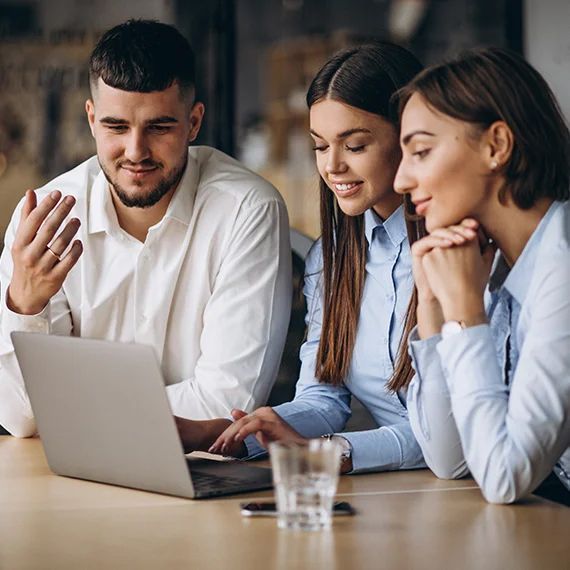 A man and two women are sitting at a table looking at a laptop computer.