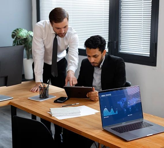 Two men are sitting at a table with a laptop and a tablet.