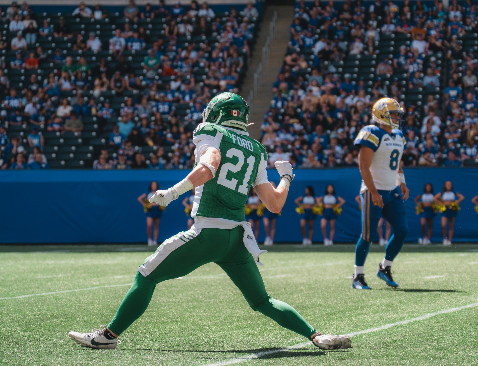 Football player in green jersey runs on field as opponent in white and blue watches nearby