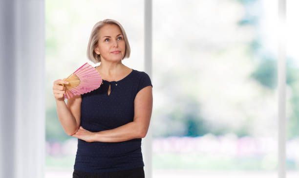 A person with short hair holds a pink hand fan in front of a blurred background.