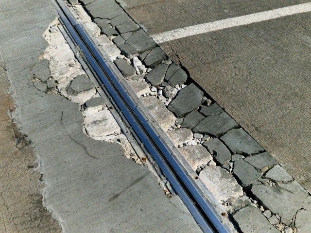 Damaged concrete barrier next to a parking space, with broken pieces and a blue metal edge.