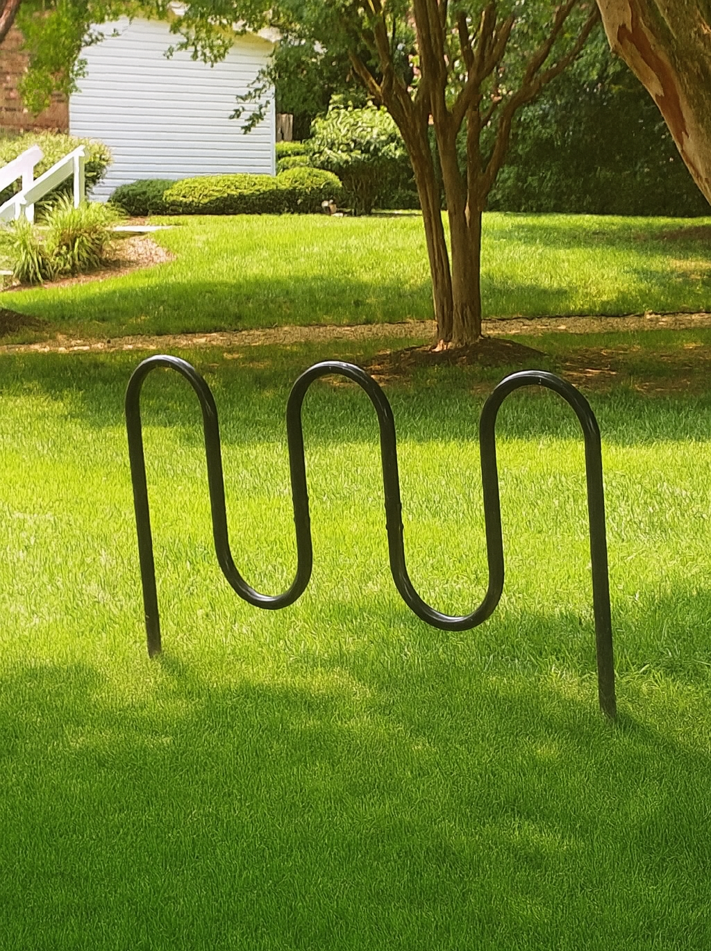 Black, wavy bike rack in a grassy yard, with a building and tree in the background.