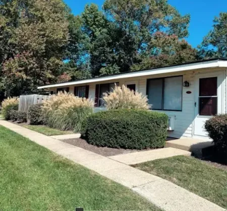 Row of white, single-story apartments with manicured landscaping, concrete walkway, and blue sky.